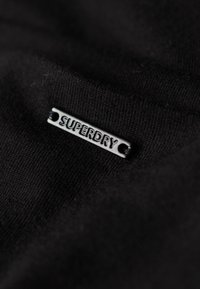 Tissu noir avec une texture en maille qui présente une étiquette rectangulaire en métal noir embossée avec "SUPERDRY". L'étiquette a deux trous pour l'attache.