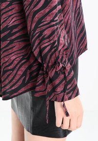 Blouse en tissu noir avec des rayures zèbres bordeaux, avec des manches froncées nouées aux poignets. Portée sur une jupe en cuir noir.