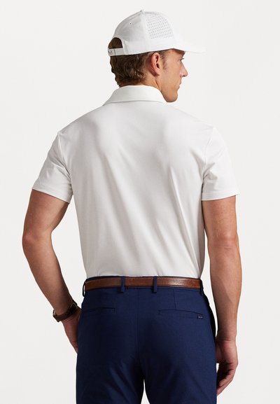 Bărbat îmbrăcat cu un tricou polo alb cu mânecă scurtă, o șapcă albă perforată, pantaloni albaștri închis cu un curea maro, văzut din spate pe un fundal simplu.