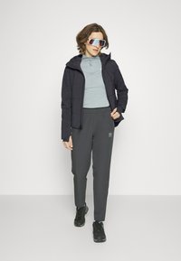 Gepolsterte schwarze Jacke über einem hellblauen Langarmoberteil, kombiniert mit grauen Sweatpants und schwarzen Turnschuhen, ergänzt mit übergroßen Sonnenbrillen.