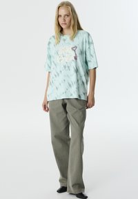 Camiseta oversized de tie-dye verde claro con un gráfico de llave colorido, combinada con pantalones grises sueltos y zapatos negros sobre un fondo liso.