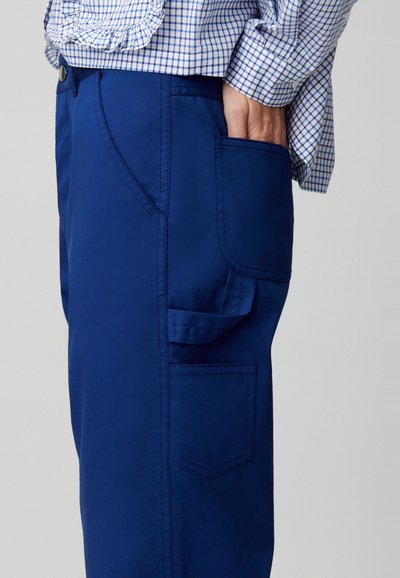 Pantaloni blu navy con una texture liscia, dotati di tasca laterale e cuciture a contrasto. Abbinati a una camicia a quadri con dettagli arricciati.