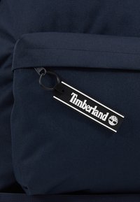 Étiquette logo Timberland en noir et blanc attachée à la fermeture éclair d'une poche de sac à dos en tissu texturé bleu marine foncé.