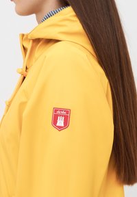 Gul vattentät jacka med huva, tillverkad i lättviktigt material och med en patch på vänster arm som visar en röd sköldlogotyp.