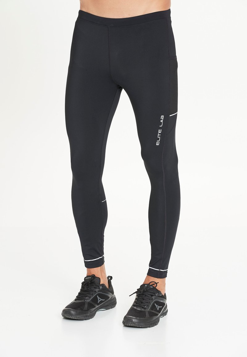 Homme portant des leggings de compression noirs avec le texte "ELITE LAB" sur la cuisse gauche et des baskets de sport noires sur fond blanc.