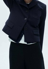 Blazer court bleu marine avec col, deux poches avant et boutons noirs. Porté sur une chemise blanche et associé à un pantalon noir.