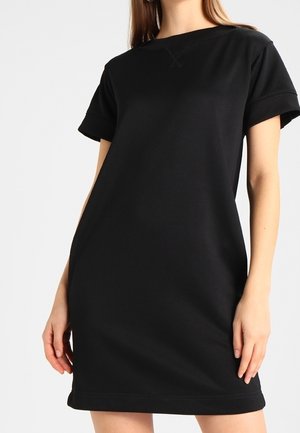 Kobieta ubrana w prostą czarną sukienkę z krótkim rękawem o luźnym kroju, stojąca na jednolitym jasnoszarym tle.