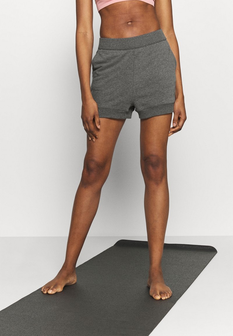 Personne debout pieds nus sur un tapis de yoga noir, portant un short de sport gris foncé et un top court rose, sur un fond clair uni.