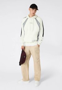 Witte hoodie met grijze en zwarte accenten, beige cargobroek en een bordeauxrode heuptas, tegen een neutrale achtergrond.