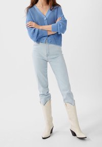 Light blue knitted cardigan with three-quarter sleeves, paired with light blue skinny jeans and cream western-style heeled boots.