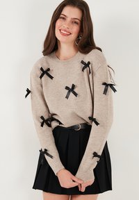Pull en maille beige avec des accents en ruban noir sur les manches, associé à une jupe plissée noire et une fine ceinture noire.