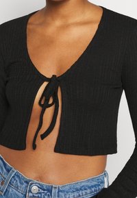 Svart ribbad kort topp med djup V-ringning, med en framknutning och lång ärmar, i kombination med högmidjade denimjeans.