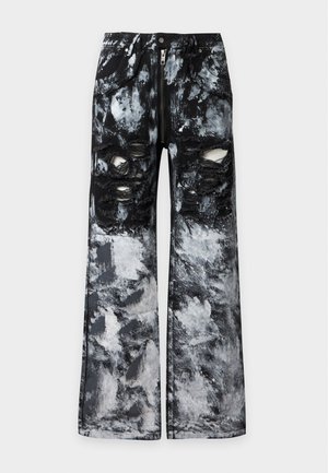 Jeune homme portant un sweat à capuche noir zippé avec des motifs et un pantalon tie-dye noir et blanc déchiré, avec des baskets slip-on à carreaux rouges et blancs.