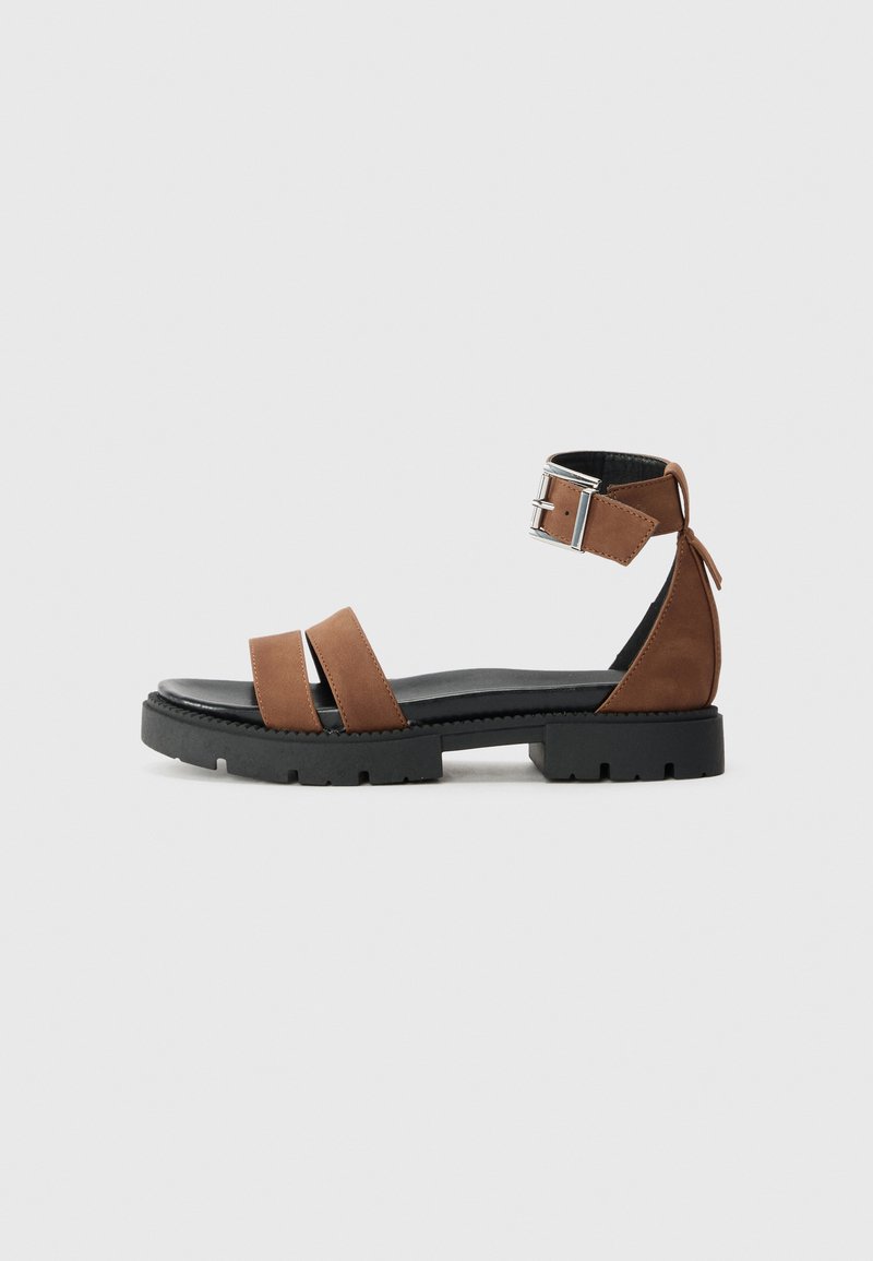 Sandales en cuir brun avec deux larges brides, une bride à la cheville avec une boucle en argent, et une semelle noire texturée. Design minimaliste.