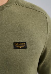 Pulover v olivno zeleni barvi z rebrastim ovratnikom, ki ima na prsih črn prenos z zlatim napisom in dizajnom kril.