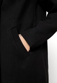 Cappotto di lana nera con tasca sul lato sinistro, dal design pulito e dalla texture morbida, che enfatizza uno stile minimalista.