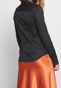 Svart silkeskjorta med långa ärmar och rundad nederkant, bärs över en orange satängkjol. Har en subtil glans och en skräddarsydd passform.