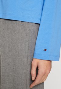Primo piano di una mano posata sul taschino dei pantaloni grigi, indossando una camicia a maniche lunghe azzurra chiara con un piccolo logo rosso, bianco e blu sul polsino.