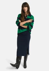 Grüner und navy gestreifter Pullover mit Kragen und Knopfakzenten, kombiniert mit einem navy Midirock mit Schlitz an der Seite und schwarzen Lederstiefeln.