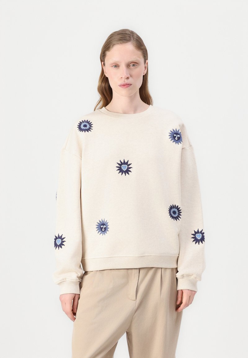 Ljusbeige sweatshirt med marinblå broderade solmotiv. Rund halsringning, lös passform och mjukt tyg. Kombinerad med ljusa byxor.