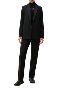 Blazer nero con risvolti in satin, abbinato a un top nero in pizzo con collo alto e pantaloni neri slim. Tessuto liscio e vestibilità sartoriale.