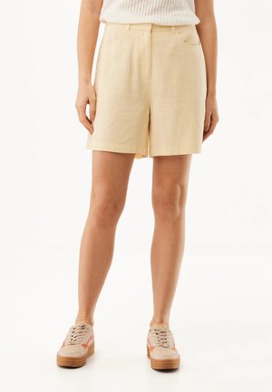 Femme portant un short en lin beige taille haute, un haut blanc sans manches, et des baskets beiges avec des accents orange, debout devant un fond blanc.