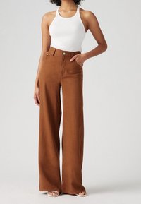 Ruske leve bukser lavet af bomuldsstof, med høj talje, forlommer og en glat tekstur. Parret med en hvid tanktop.