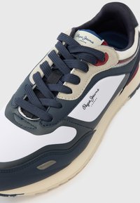 Αθλητικό παπούτσι σε ναυτικό μπλε, λευκό και μπεζ με λείες επιφάνειες δέρματος και υφές από πλέγμα. Διαθέτει χοντρές, σκούρες κορδόνια και λογότυπο στο πλάι.