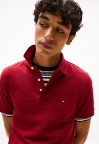 Rdeča polo majica z teksturirano tkanino, z ovratnikom s tremi gumbi, črtastim notranjim podlogom in majhno majico na prsih.