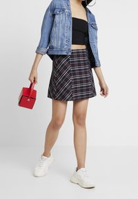 Veste en denim, crop top noir, jupe à carreaux en marine, rouge et blanc, sac à main rouge avec une poignée, et baskets blanches. Design de jupe courte en A.