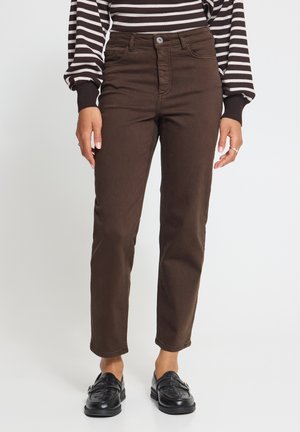 Personne portant un pantalon droit marron, des mocassins tissés noirs et une chemise rayée noire et blanche à manches longues, debout sur un fond blanc.