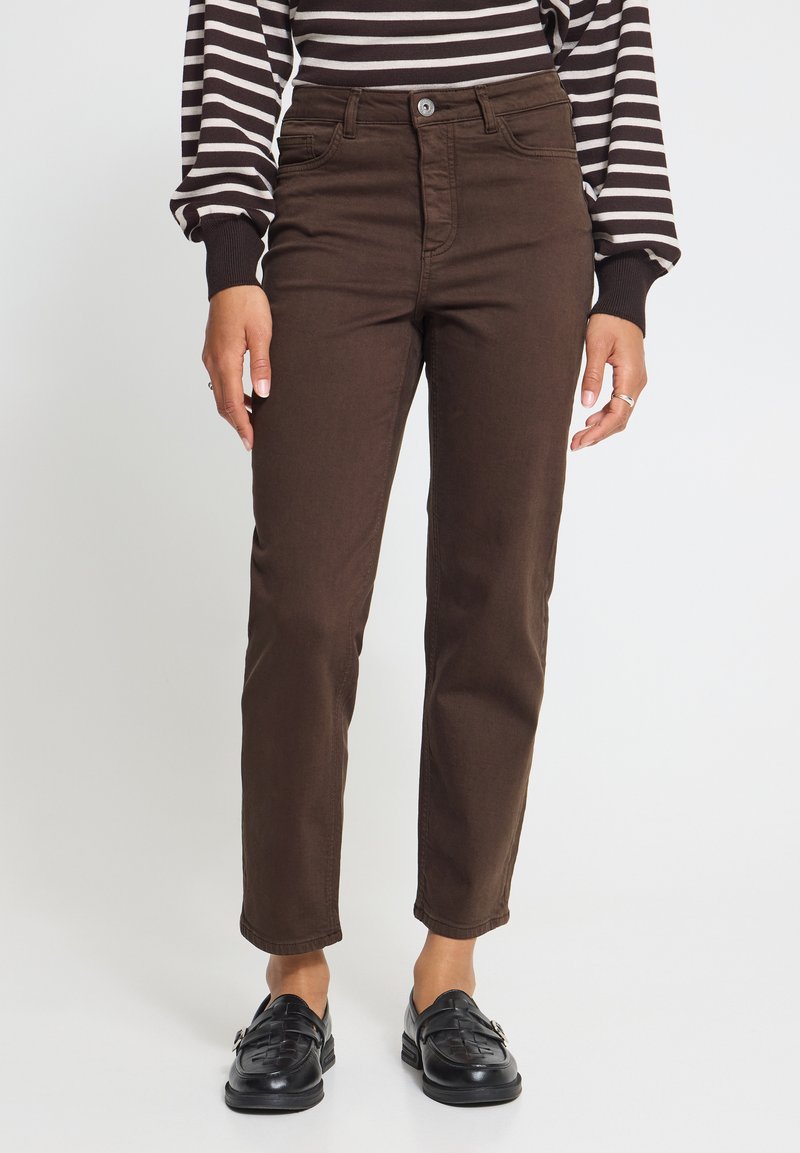 Personne portant un pantalon droit marron, des mocassins tissés noirs et une chemise rayée noire et blanche à manches longues, debout sur un fond blanc.