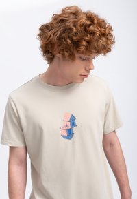 Beige katoenen t-shirt met een grafisch ontwerp van twee gestapelde huizen in roze en blauw met een lichte textuur, gepresenteerd op een model.