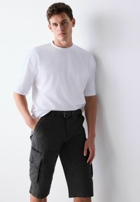 Homme portant une chemise blanche à manches courtes et un short cargo noir avec des poches latérales, debout contre un mur blanc avec les mains dans les poches.
