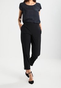 Černé kalhoty na míru s hladkou texturou a uvolněným střihem; v kombinaci s tmavým, vzorovaným tričkem s krátkým rukávem. Černé boty s pásky na kotnících.