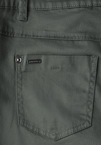 Dunkelgrünes, rechteckiges Stofftasche mit feiner Textur und einem Markenmetallanhänger, der mit einem Knopfdetail befestigt ist.