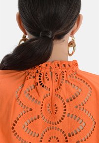 Oranje blouse met intrigerende kantdetails op de achterkant, met cirkelvormige patronen en een sleutelgatopening bij de hals. Soepele textuur.
