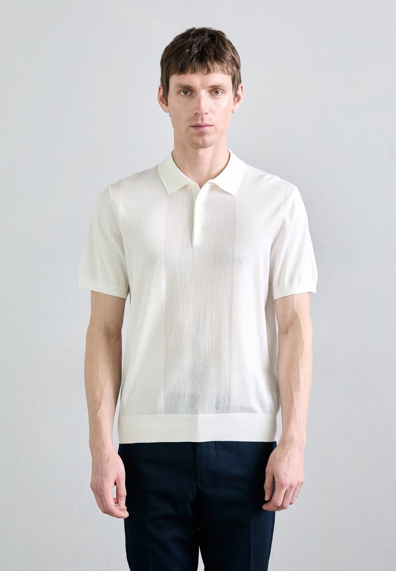 Kortärmad vit pikétröja med texturerad vertikal panel, krage och smal passform, kombinerad med mörka byxor.