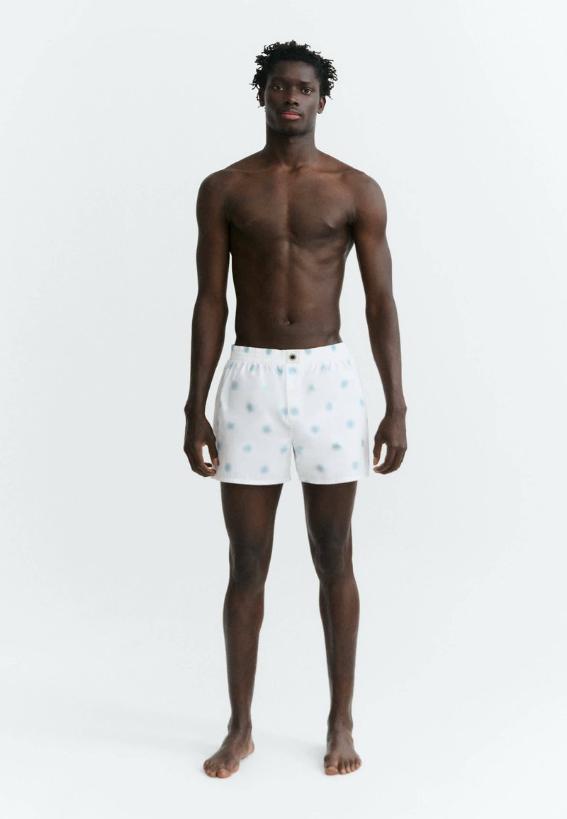 Homme grand, torse nu, peau foncée, portant un boxer blanc à pois bleu clair, debout pieds nus sur un fond blanc uni.