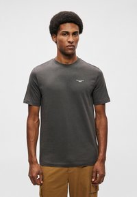 Tumši pelēks īsām piedurknēm T-krekls, kas izgatavots no kokvilnas, ar apaļu kakla izgriezumu. Tam ir neliels balts logotips kreisajā krūtī.