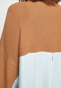 Šedohnědý oversized pulovr, který se vyznačuje texturovaným pleteným vrškem a hladkým spodním dílem se jemným švem na přechodu.