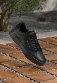 Adidași negri Tommy Hilfiger, cu șireturi, pe plăci de lemn în aer liber, cu un copac mic verde și un zid de beton în fundal.
