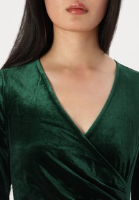 Žalias aksominis apvyniojamas viršus su V formos kaklo iškirpte, lygia tekstūra ir ilgomis rankovėmis. Įrengtas drapiruojant per krūtinę, kad būtų pritaikytas stilius.