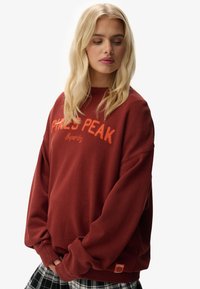 Sudadera burdeos con bordado "PHLES PEAK" en naranja, corte holgado, puños acanalados y pequeña etiqueta de logo en la parte inferior izquierda. Textura suave.