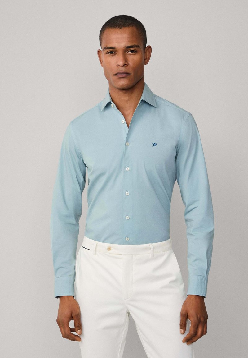 Lichtblauw lange mouwbutton-up overhemd met een kraag, voorzien van een klein marineblauw logo op de borst, gedragen met witte broek.