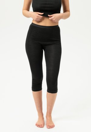 Personne portant des leggings courts noirs et un haut noir sans manches, debout pieds nus sur un fond clair uni.
