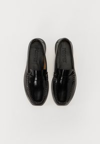Mocassins en cuir noir avec des coutures décoratives, texture lisse et léger éclat. L'intérieur présente le nom de la marque embossé sur la semelle intérieure.