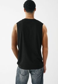 T-shirt sans manches noir en coton avec une coupe décontractée, présentant un col rond et des coutures d'épaule cousues. Porté avec un jean en denim délavé.