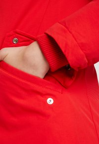 Rode jas met een voorzak met drukknoopsluiting en geribbelde manchetten. Gladde textuur en moderne designelementen zijn zichtbaar.