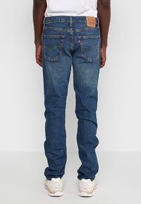 Homme portant un jean Levi's bleu et des baskets blanches, debout dos à la caméra devant un fond uni.